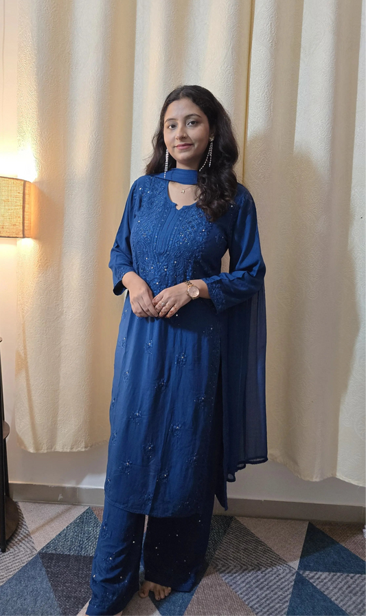 Woman in a blue traditional outfit standing in a room with a lamp and curtains.