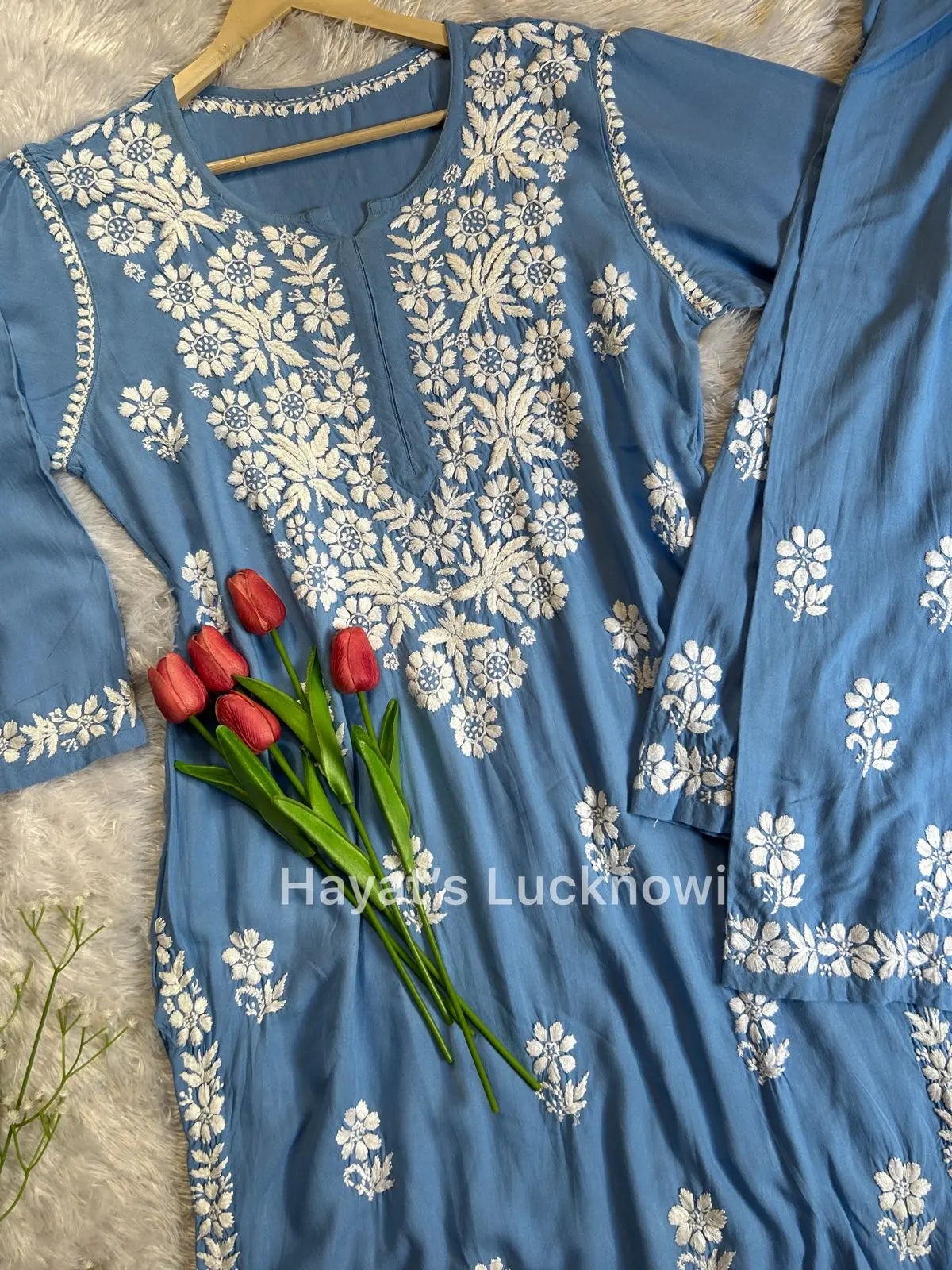 Powder blue chikankari rayon kurta set with intricate white floral embroidery, displayed on a white fur background with decorative red rose petals and artificial tulips. The set is from Hayat&